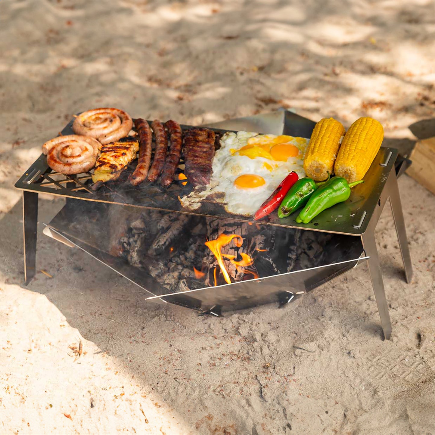 Die FENNEK Feuerschale Hexagon steht am Strand am See. Darüber ist der FENNEK 4fire aufgestellt, auf welchem 1 Steak, 3 Merguez, 2 Bratwurstschnecken, 1 Hähnchenbrustfilet, 2 Spiegeleier, 1 rote Paprika, 2 grüne Paprika und 2 Maiskolben gegrillt wird. 
