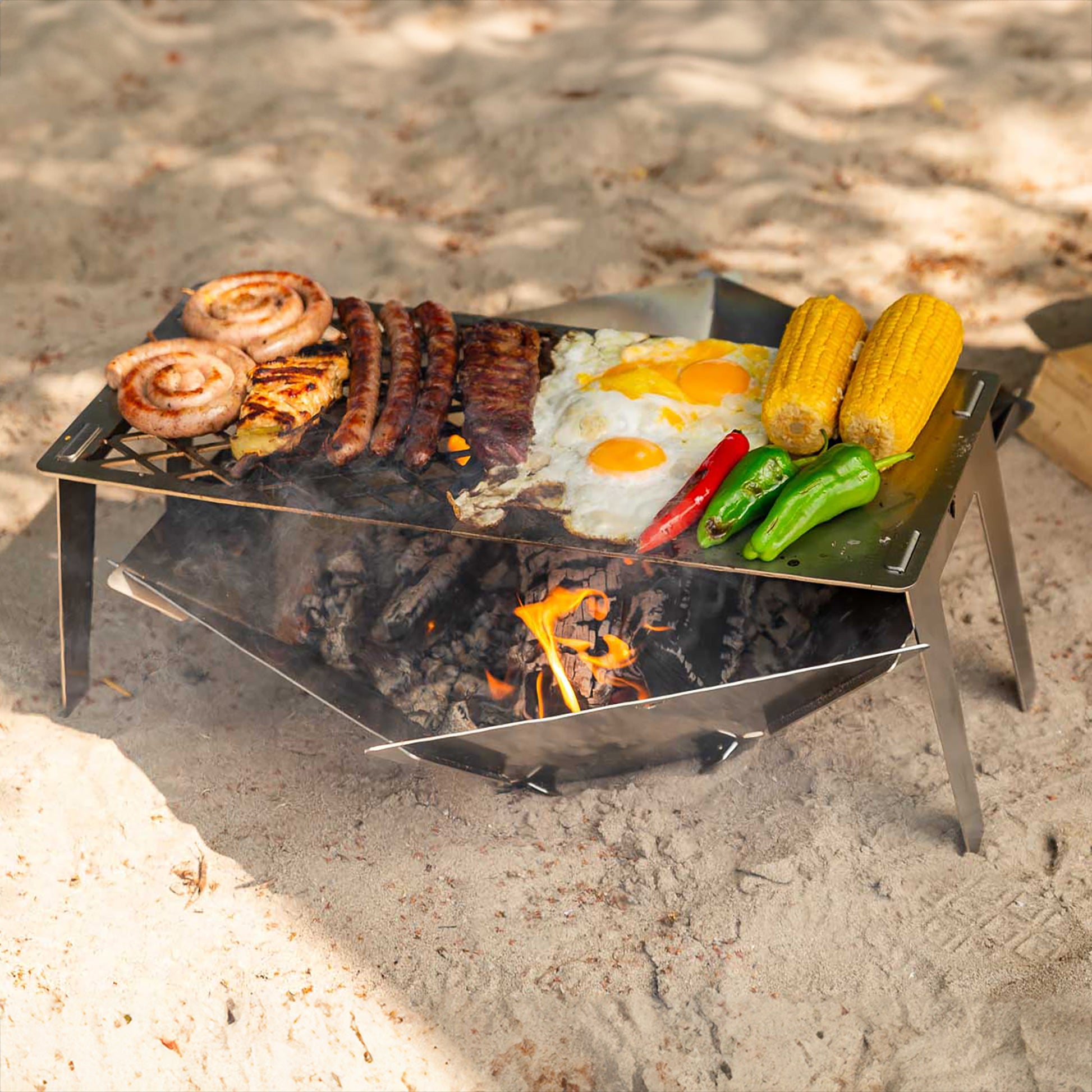 Die FENNEK Feuerschale Hexagon steht am Strand am See. Darüber ist der FENNEK 4fire aufgestellt, auf welchem 1 Steak, 3 Merguez, 2 Bratwurstschnecken, 1 Hähnchenbrustfilet, 2 Spiegeleier, 1 rote Paprika, 2 grüne Paprika und 2 Maiskolben gegrillt wird. 