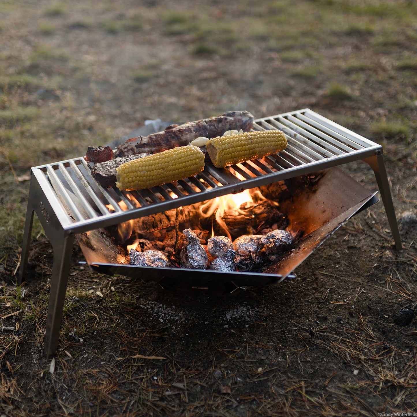 Der FENNEK Klapprost XL steht über der FENNEK Feuerschale Hexagon in welcher glühende Kohle und Kartoffel in Alufolie liegen Alles steht auf grünem Gras.