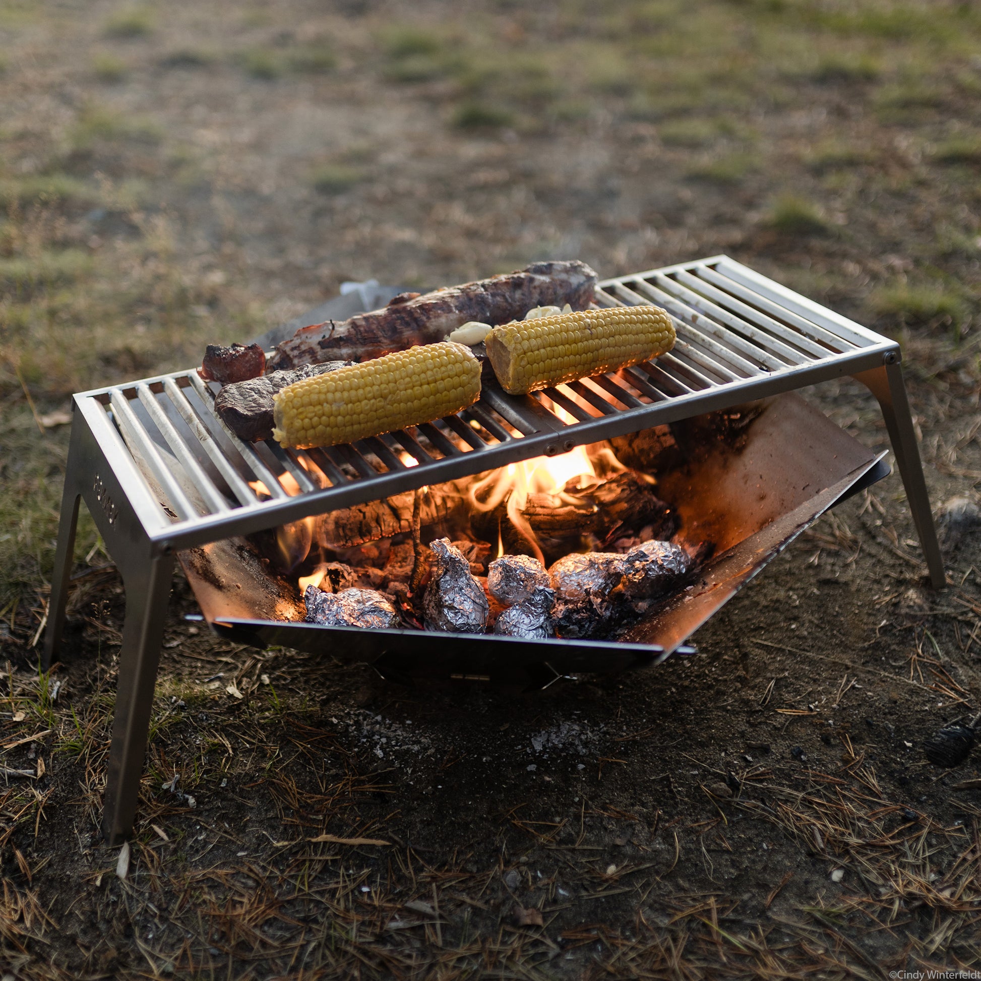 Der FENNEK Klapprost XL steht über der FENNEK Feuerschale Hexagon in welcher glühende Kohle und Kartoffel in Alufolie liegen Alles steht auf grünem Gras.