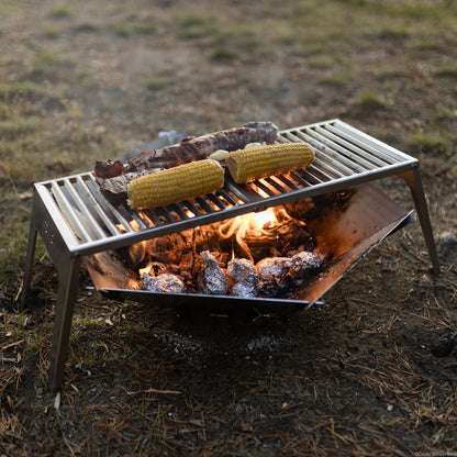 Der FENNEK Klapprost XL steht über der FENNEK Feuerschale Hexagon in welcher glühende Kohle und Kartoffel in Alufolie liegen Alles steht auf grünem Gras.