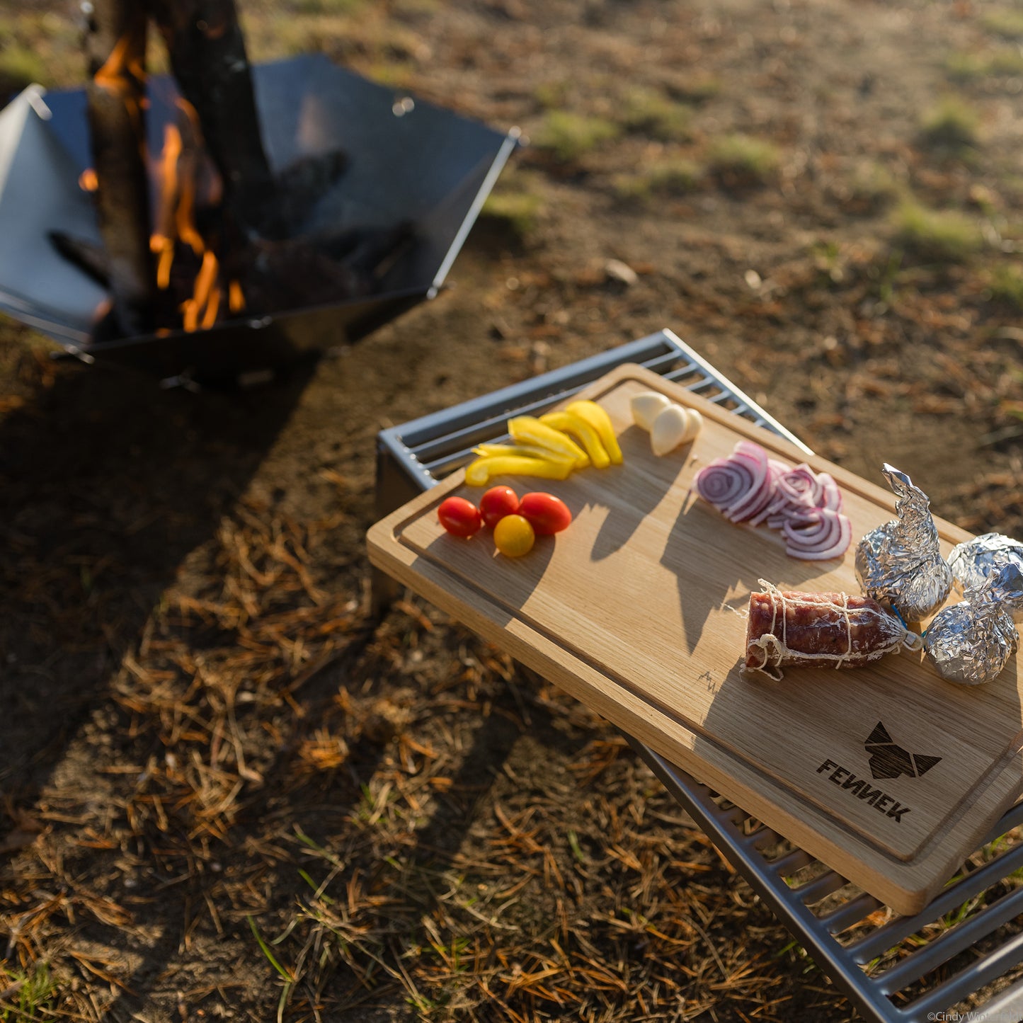 Auf einem FENNEK Klapprost XL liegt ein FENNEK Schneidebrett, das mit etwas Gemüse und Wurst beladen ist. Die FENNEK Feuerschale Hexagon ist im Hintergrund zu sehen, in ihr brennt Feuer. 