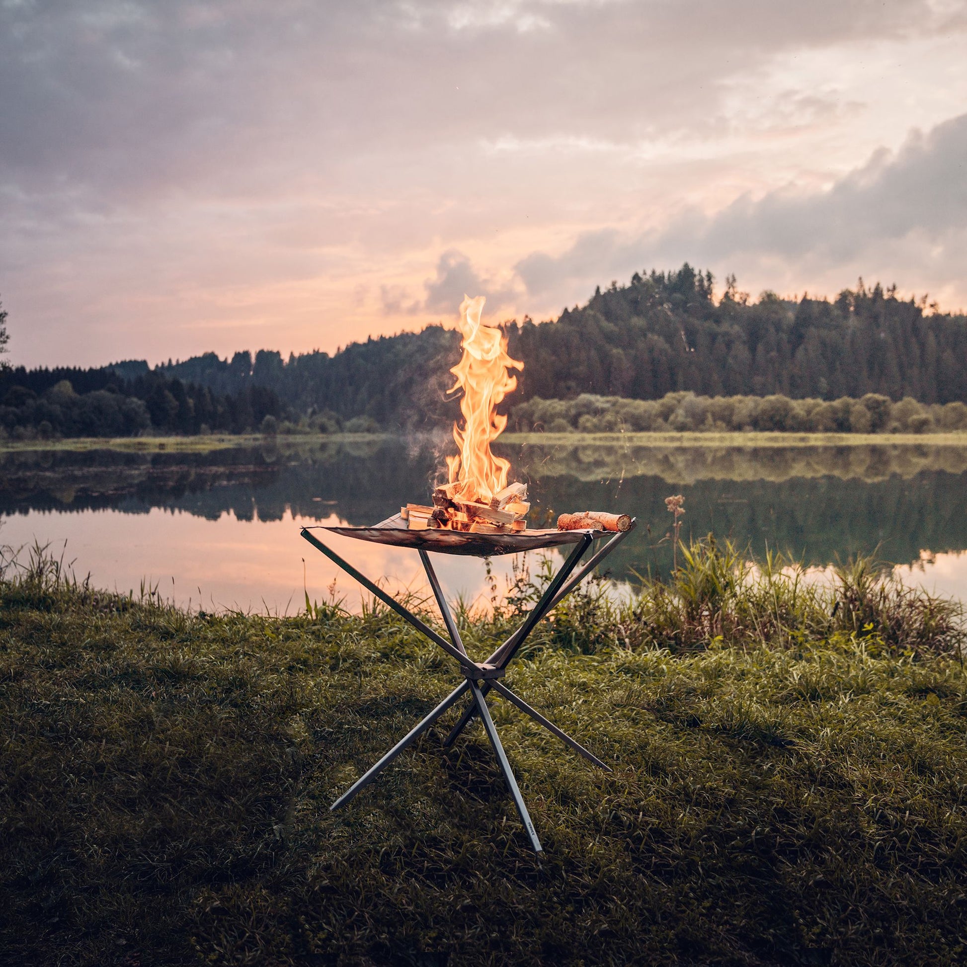 Das in der Natur aufgestellte Feuernetz im Einsatz mit brennendem Holz darauf und einer stimmungsvollen Aussicht auf ein stilles Gewässer mit Spiegelung der dichtbewachsenen Landschaft im Hintergrund.
