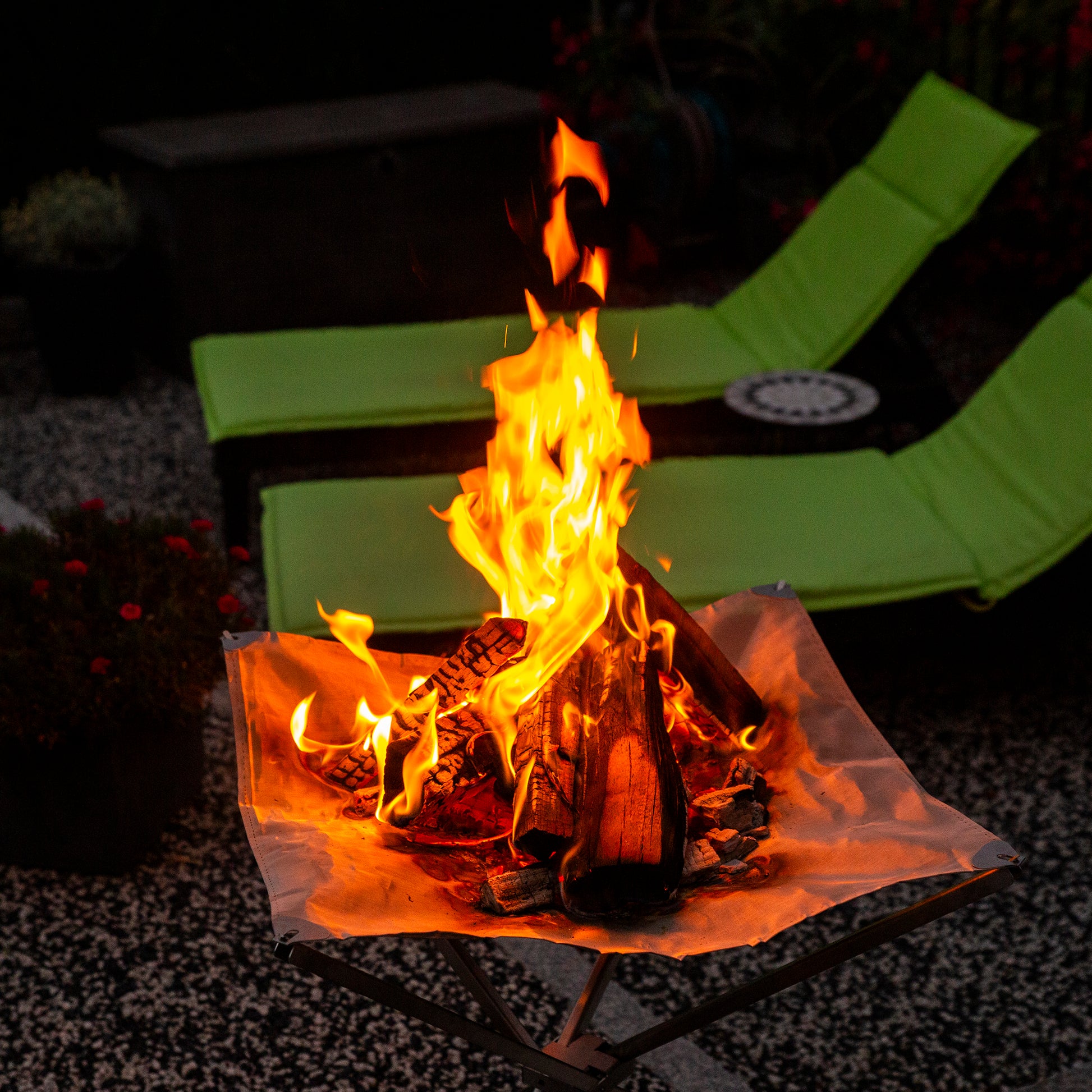 Brennendes Feuer im Feuernetz bei Dunkelheit, das im Garten steht und im Hintergrund zwei Sonnenliegen.