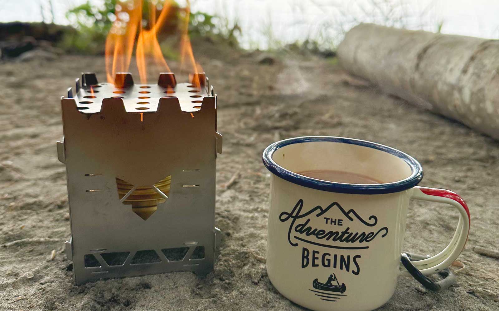 Der FENNEK Hobokocher Solo aus Edelstahl steht auf einem sandigen Untergrund am See. Daneben steht eine Tasse mit einem Heißgetränk.