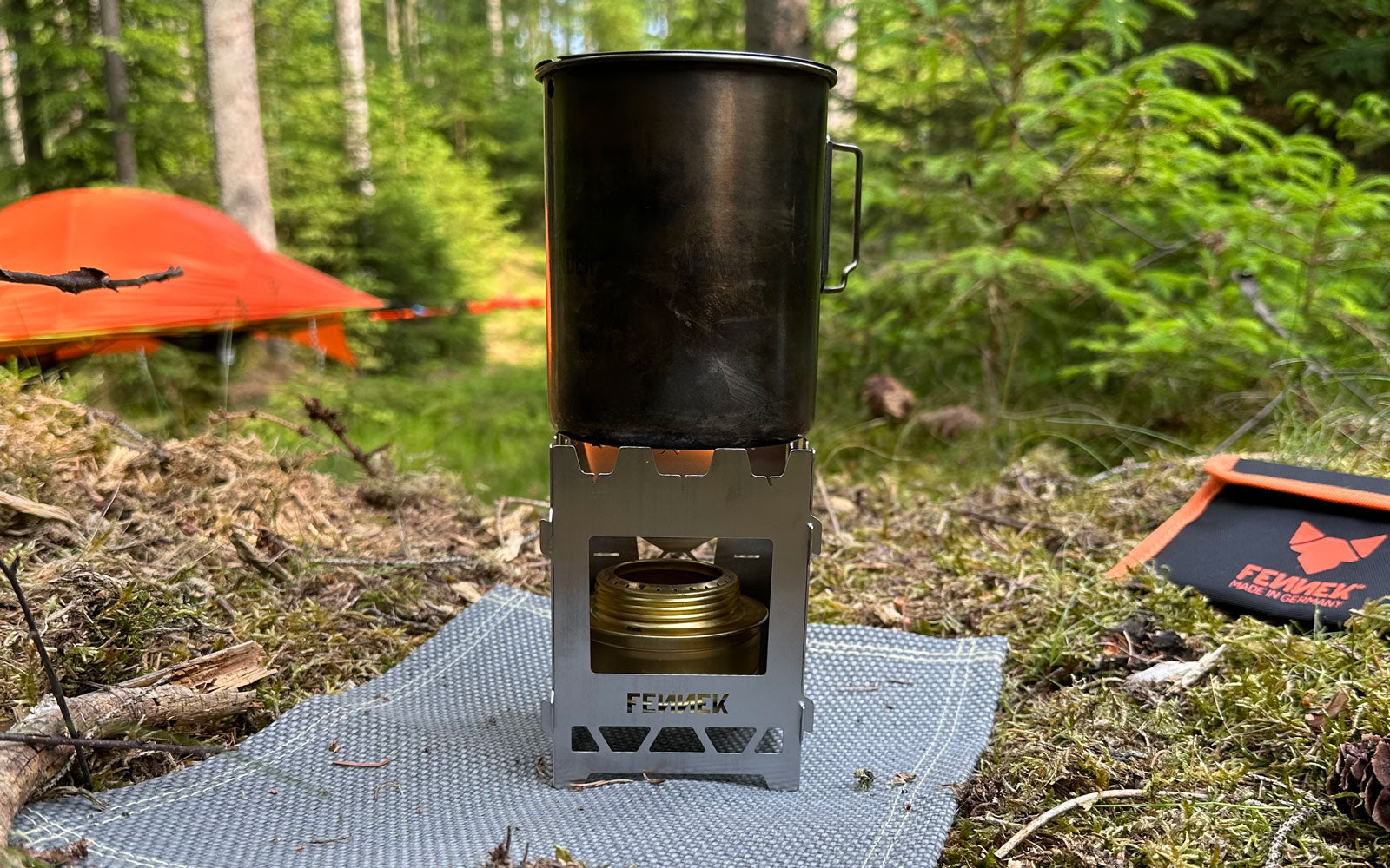 Der FENNEK Hobo Solo aus Edelstahl im Wald in Szene gesetzt. Er steht auf der FENNEK Flammschutzmatte und befeuert einen Topf. Im Hintergrund liegt die dazugehörige Tasche.
