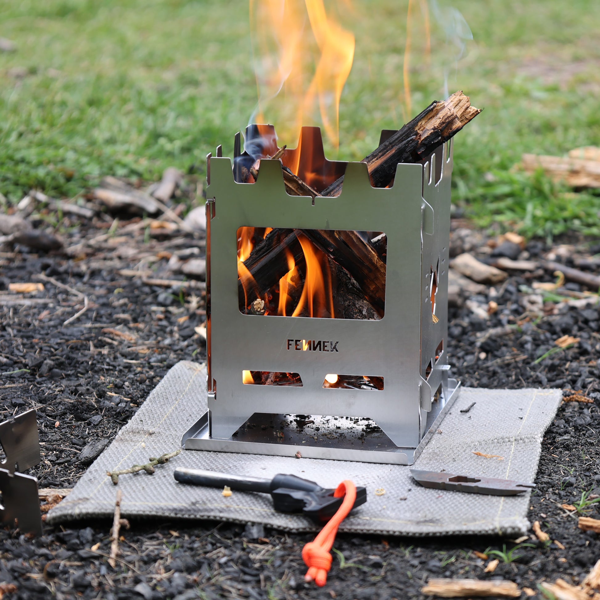 Edelstahl Hobokocher auf einer Hitzschutzmatte in der Natur im Einsatz mit Blick durch die Frontöffnung auf das brennenden Holz, davor liegt ein Feuerstahl