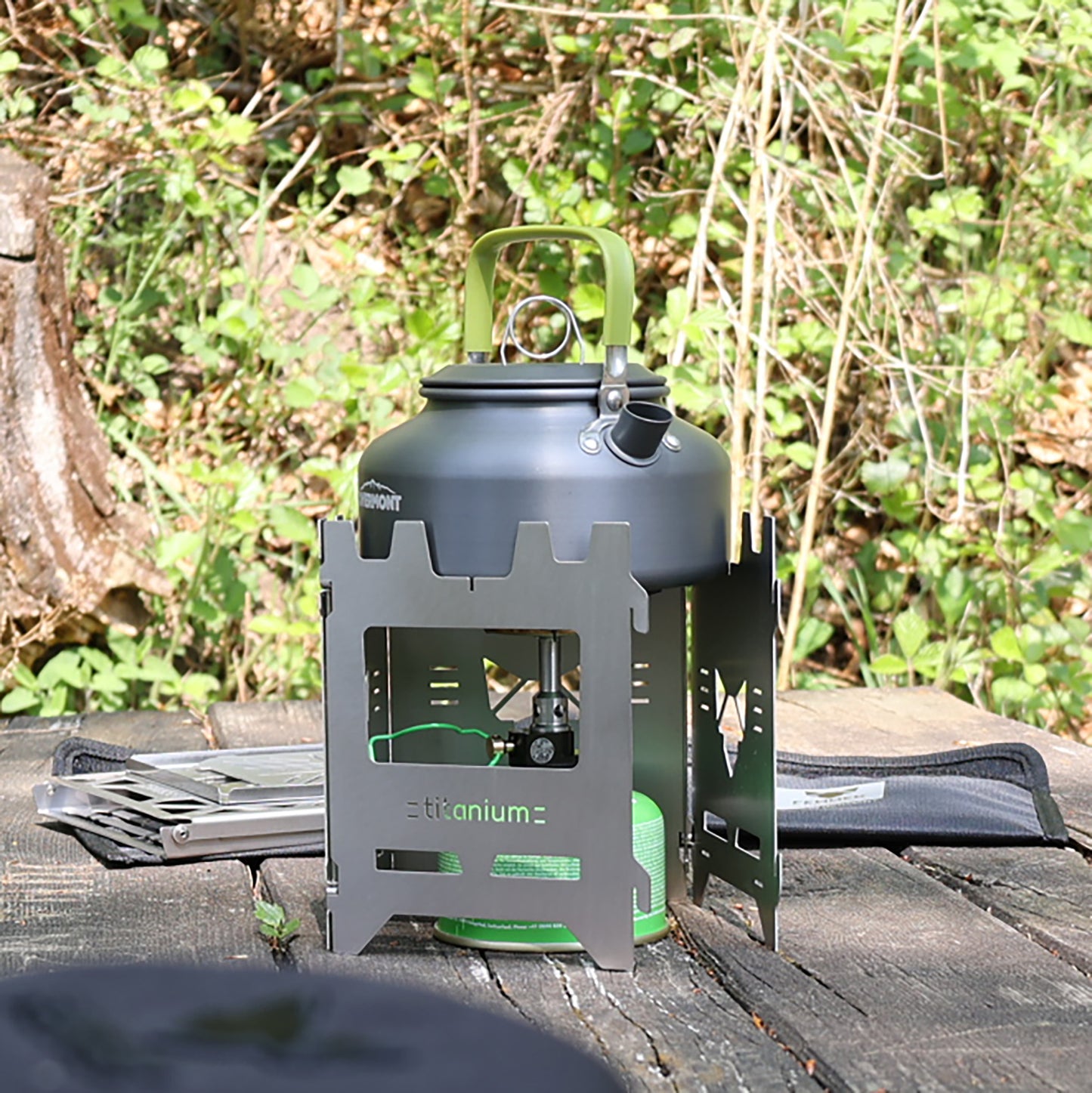 Hobokocher am Tisch einer Sitzgruppe im Wald, nicht ganz geschlossen, als Windschutz um einen Gaskocher gestellt, auf dem ein Wasserkessel zum kochen gebracht wird.