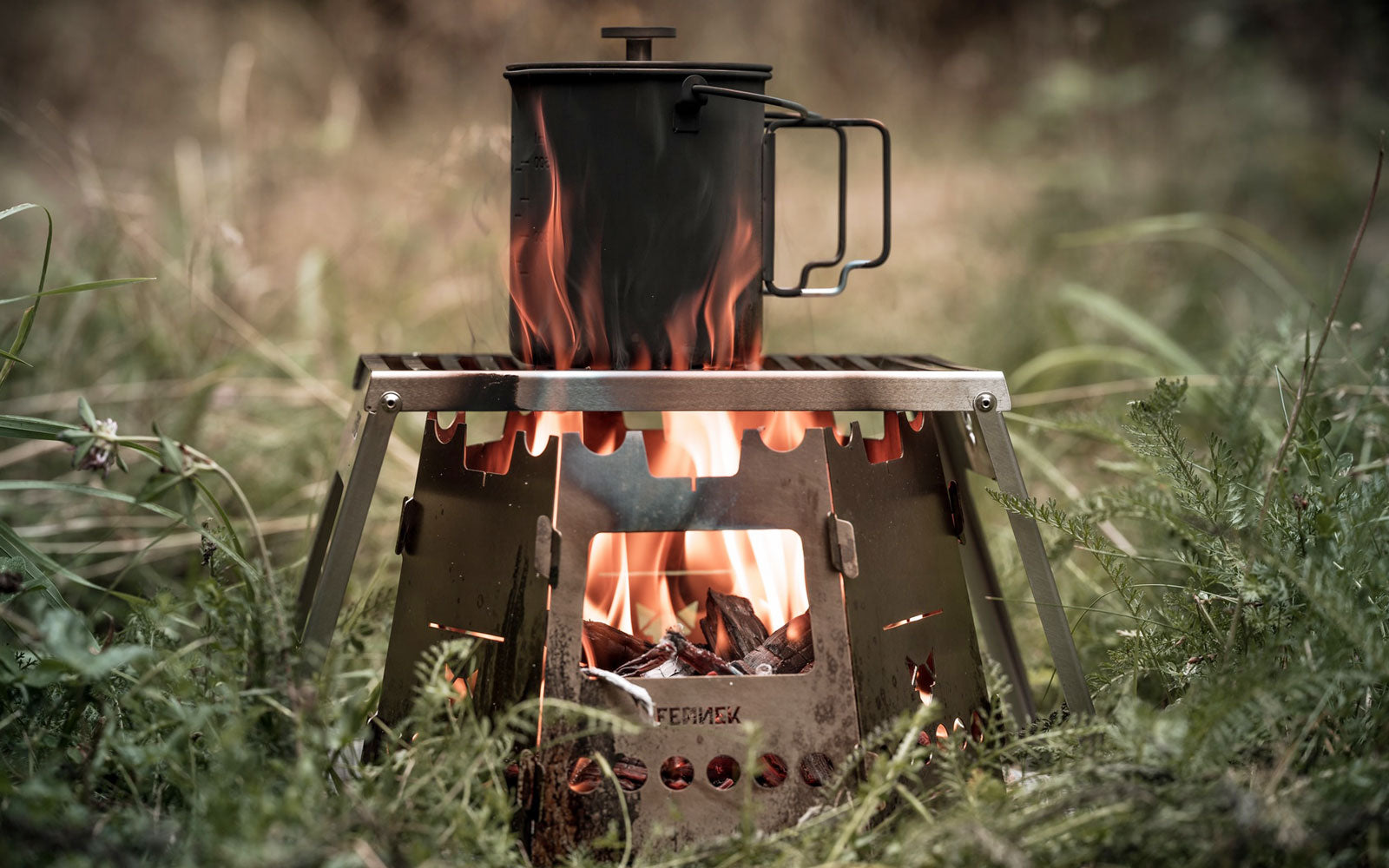 Der FENNEK Hobokocher mit Erweiterung steht auf einer Wiese. Darüber ist der Klapprost, auf welchem ein Topf über der Flamme kocht.