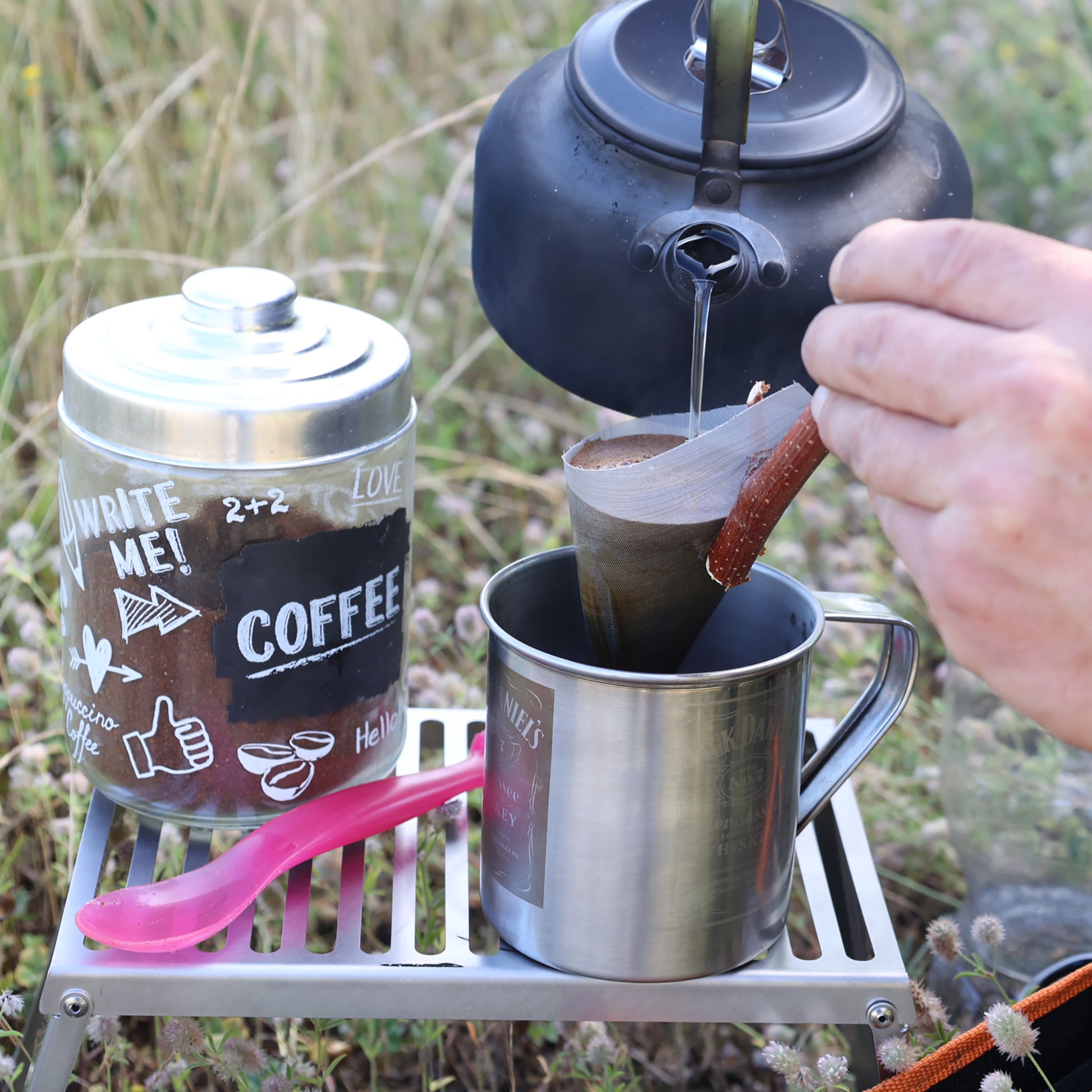 Kaffee wird mit heißem Wasser aus einem schwarzen Wasserkessel in einem Kaffeefilter aufgegossen der in eine Edelstahltasse gehalten wird die, wie auch der Behälter mit dem Kaffeepulver auf dem Klapprost abgestellt ist.