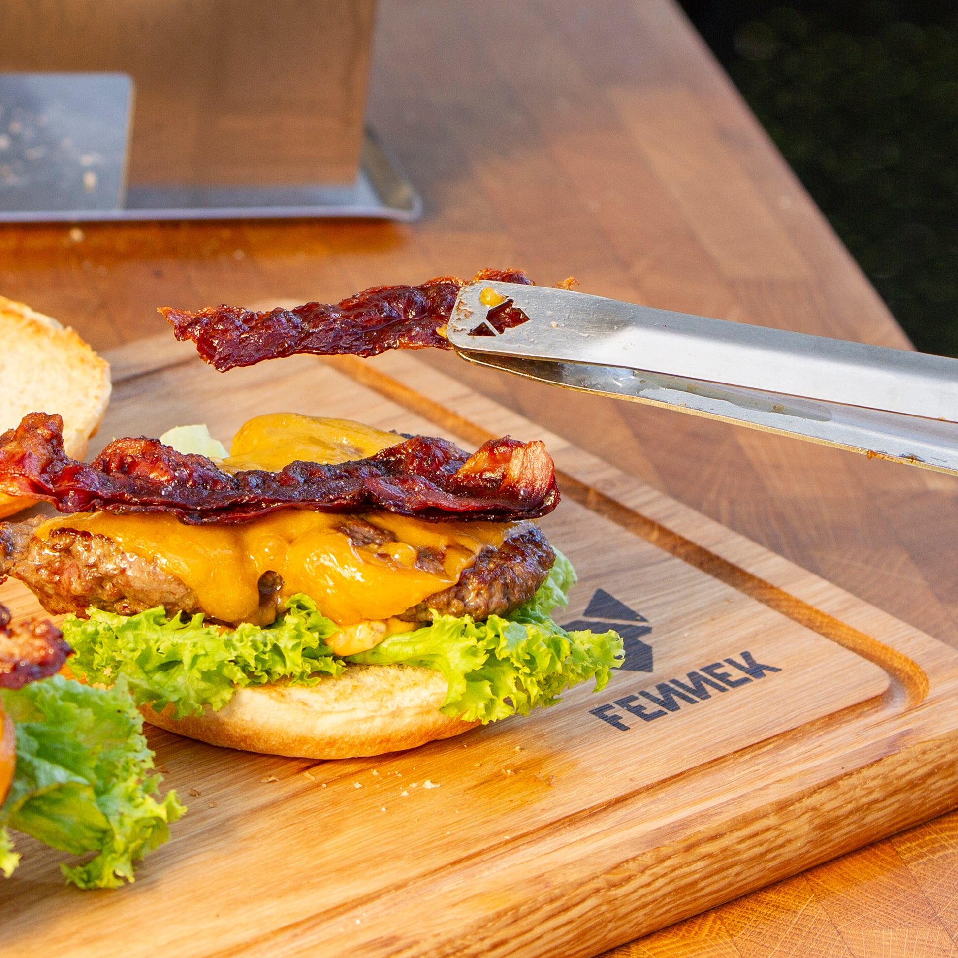 Mit Grillzange wird ein Stück knusprig gebratener Bacon auf einen Hamburger gelegt.