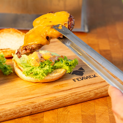 Mit Käse überbackenes Hamburger Patty wird mit Grillzange aus Edelstahl auf ein Brötchen gelegt, darunter ein Holzbrett mit Fennek Logo-Branding.