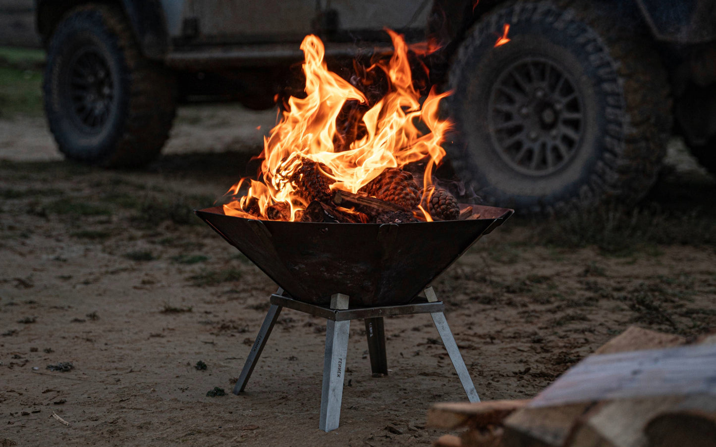 Die FENNEK Feuerschale Oktagon mit den optionalen Füßen steht vor einem Land Rover am Strand. Es ist Abendstimmung. Ein schönes Feuer lodert in der Feuerschale.