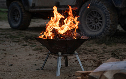 Die FENNEK Feuerschale Oktagon mit den optionalen Füßen steht vor einem Land Rover am Strand. Es ist Abendstimmung. Ein schönes Feuer lodert in der Feuerschale.