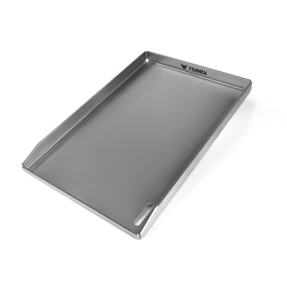 Freisteller der FENNEK Plancha 45 x 30 x 3,5 cm. Sie hat eine hochgezogene Vorderkante, damit kein Grillgut herunterfallen kann. Zusätzlich hat sie vorne rechts in der Ecke ein großzügiges Ablaufloch, worüber überschüssiges Fett und Grillreste in die Fettauffangwanne entsorgt werden können. Die vier Ecken sind offen, damit hier auch das überschüssige Fett abgeleitet werden kann. Ein Hingucker ist der eingravierte FENNEK Schriftzug.