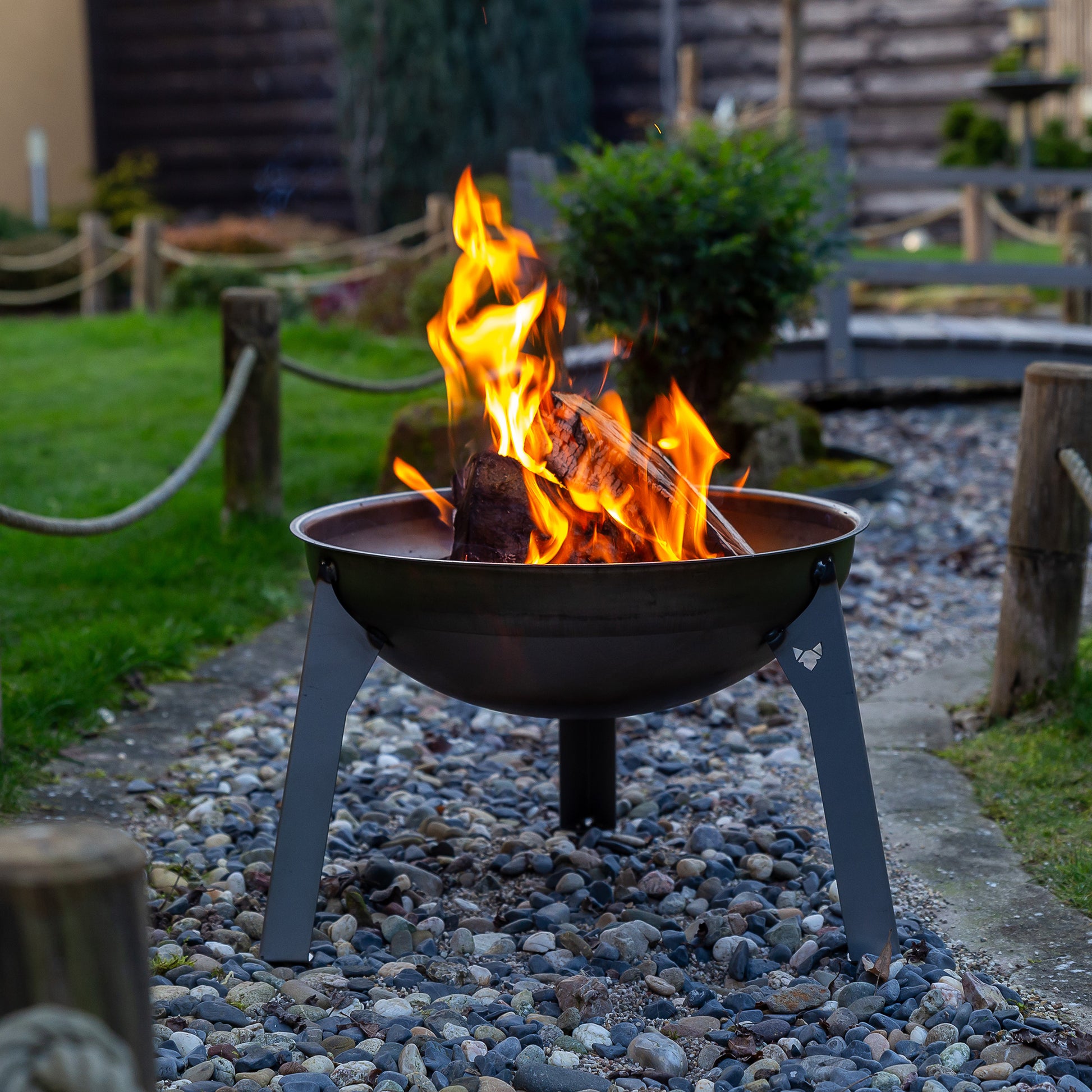 Feuerschale Rustic mit loderndem Feuer auf Kiesbett in einem schön angelegten Garten.