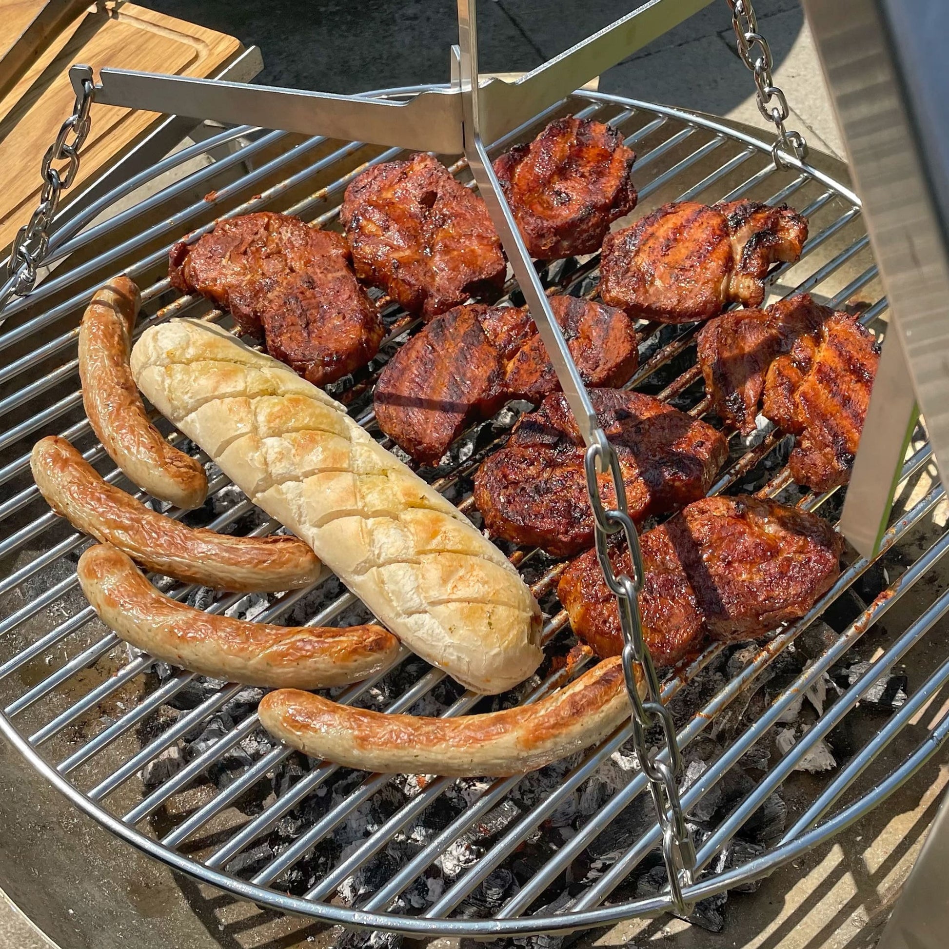 saftige Steaks und Bratwürste sowie ein Knoblauchbaguette auf der FENNEK Schwenkgrill Erweiterung der FENNEK Feuerschale Allround. Von der Seite fotografiert.