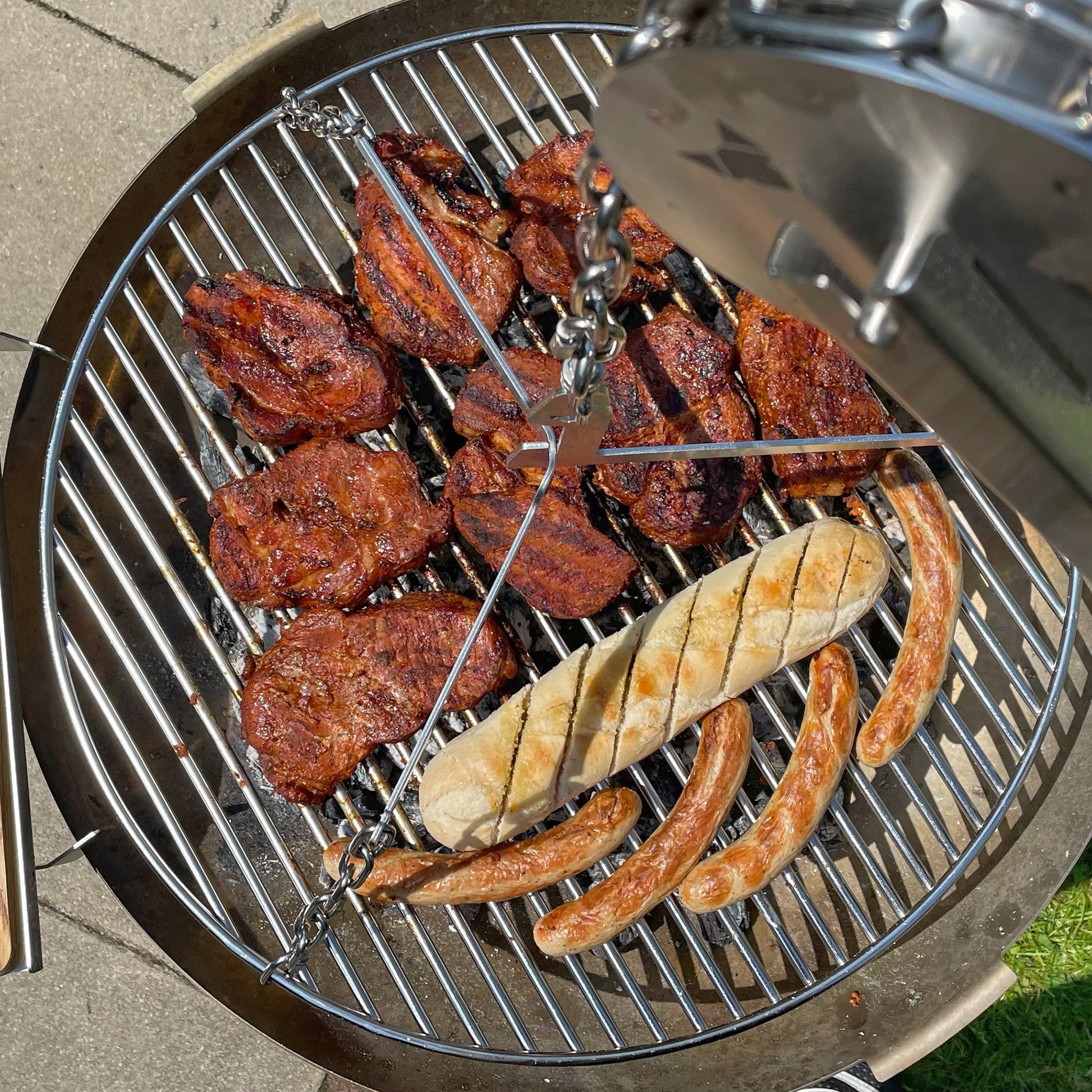 Blick von oben auf den Schwenkgrill und dem Grillrost mit Grillfleisch, Baguettebrot und Bratwürsten darauf.