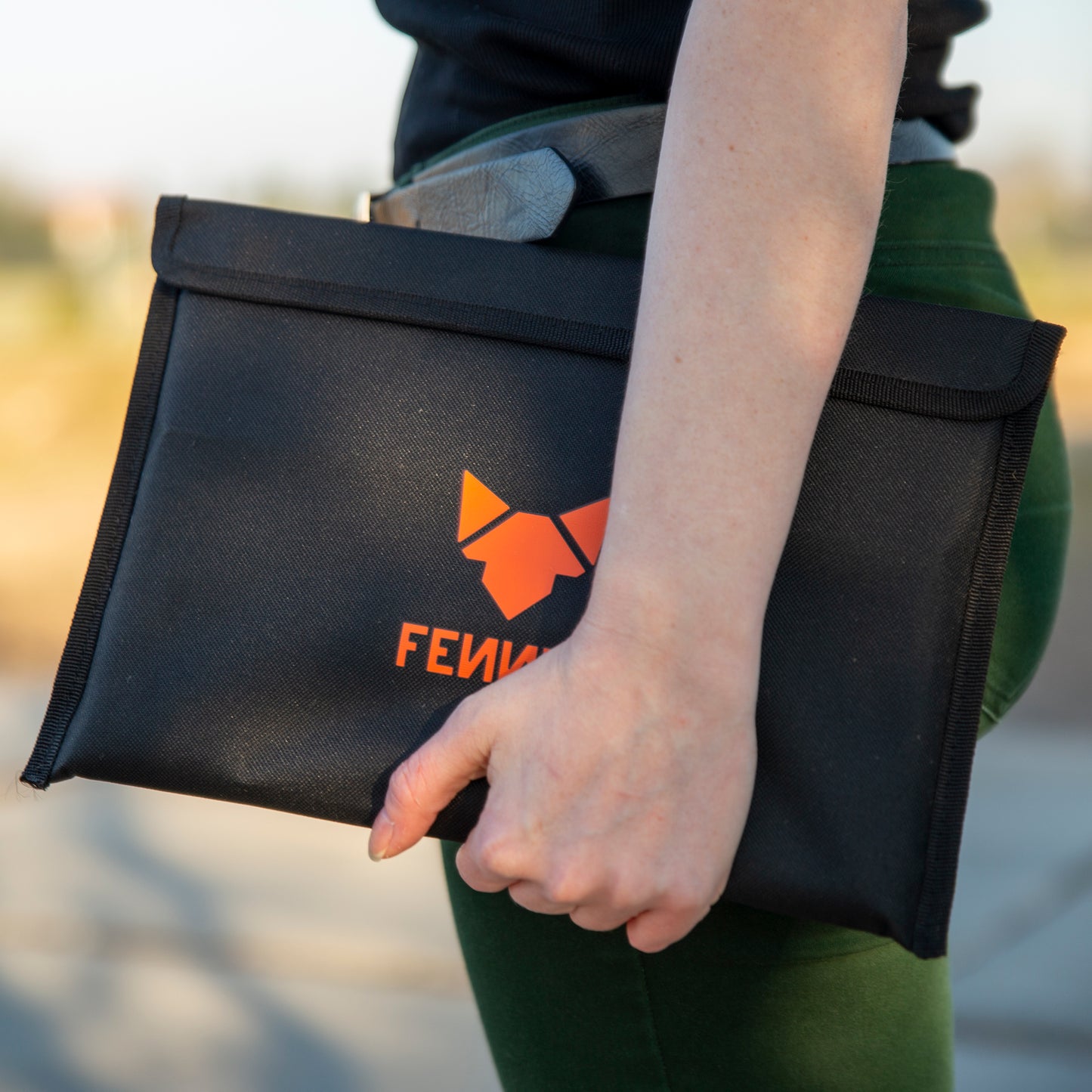 Eine Person trägt eine grüne Hose und hält auf Hüfthöhe eine schwarze Nylon-Tasche mit FENNEK Logo-Aufdruck