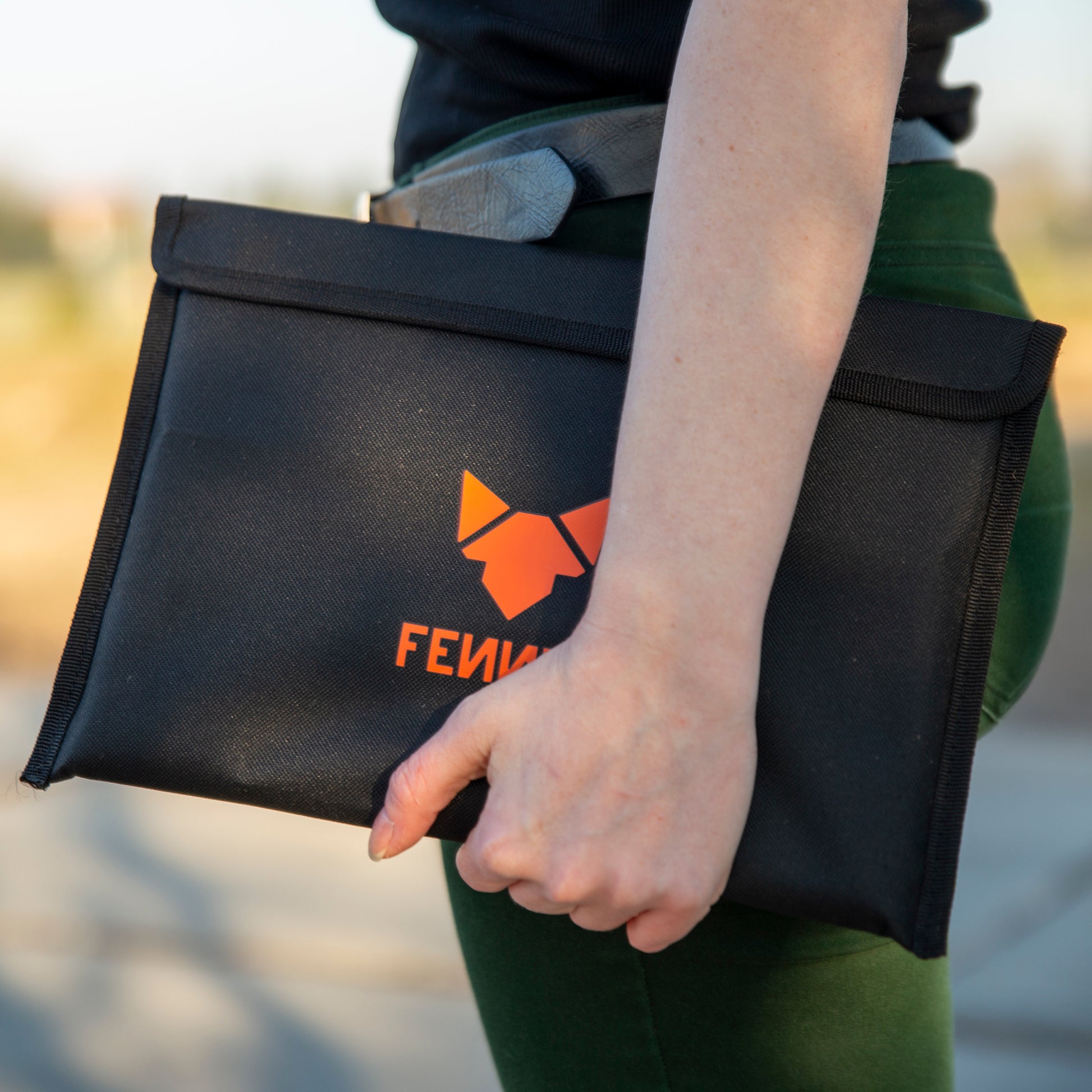 Eine Person trägt eine grüne Hose und hält auf Hüfthöhe eine schwarze Nylon-Tasche mit FENNEK Logo-Aufdruck