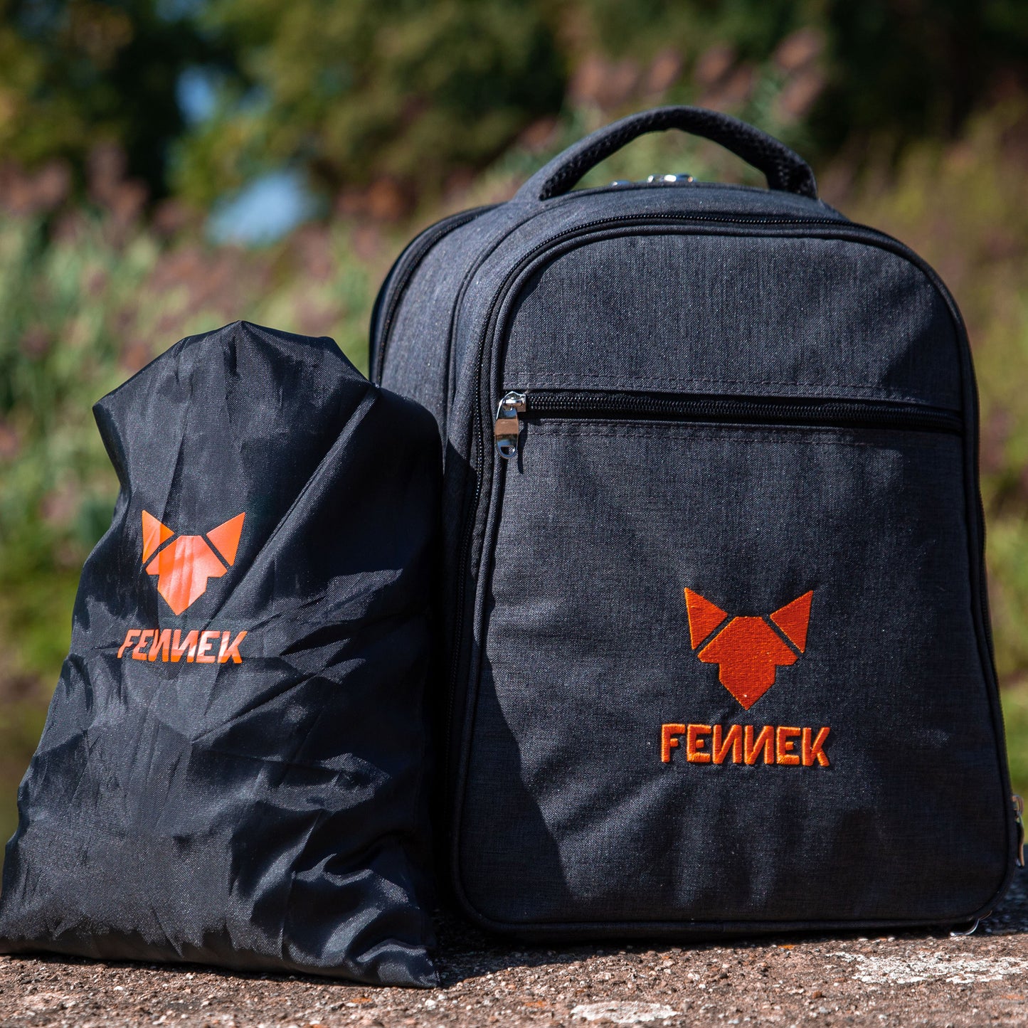 Schwarzer Kohlebeutel aus Nylon und daneben Rucksack mit FENNEK Logo-Applikation stehend auf dem Boden im Hintergrund unscharf grüne Natur.