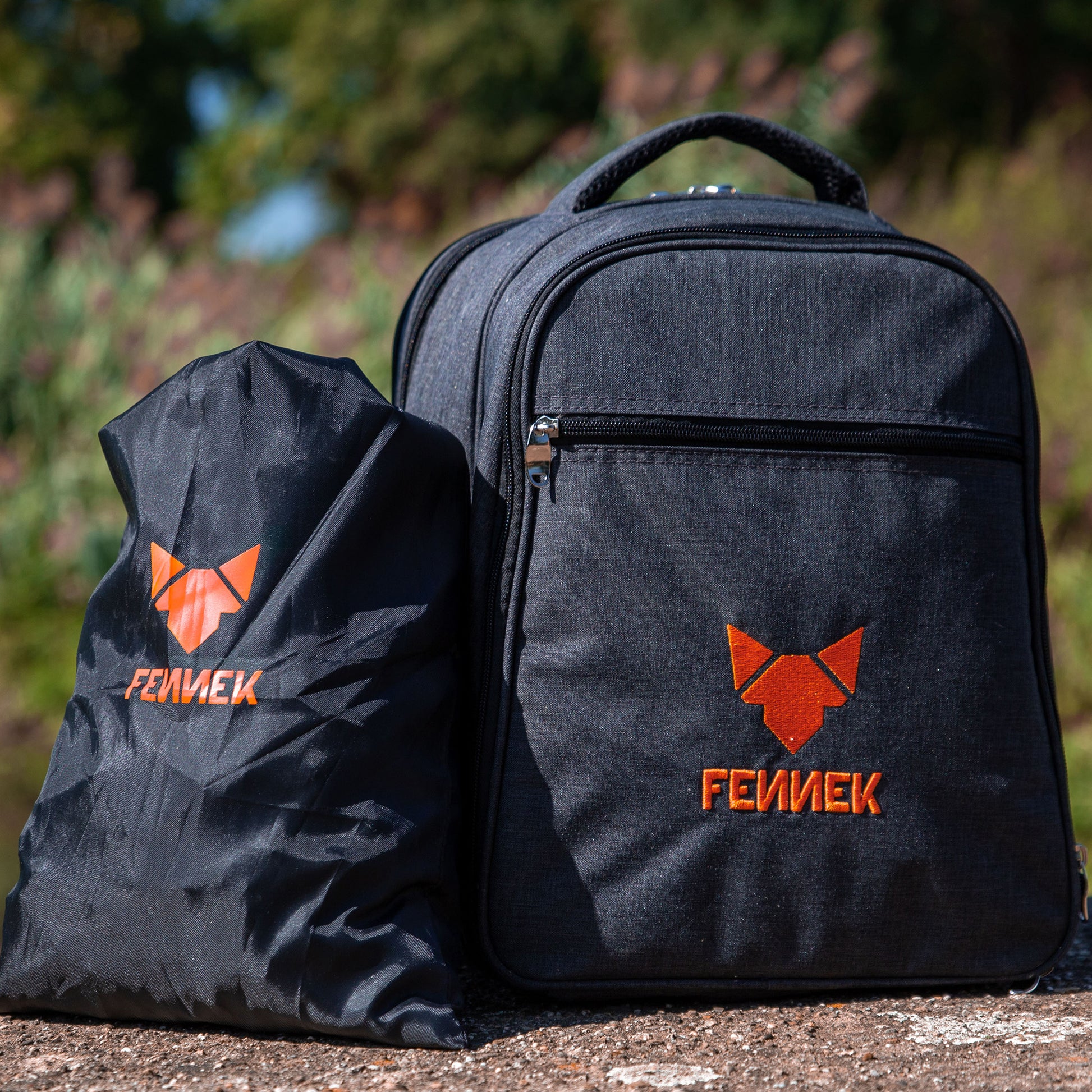 Schwarzer Kohlebeutel aus Nylon und daneben Rucksack mit FENNEK Logo-Applikation stehend auf dem Boden im Hintergrund unscharf grüne Natur.