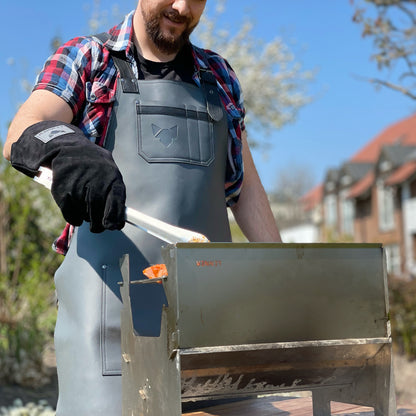 Schwarzer Grillhandschuh beim Grillen im Einsatz, getragen von einem Mann mit Ledergrillschürze, hält eine Grillzange