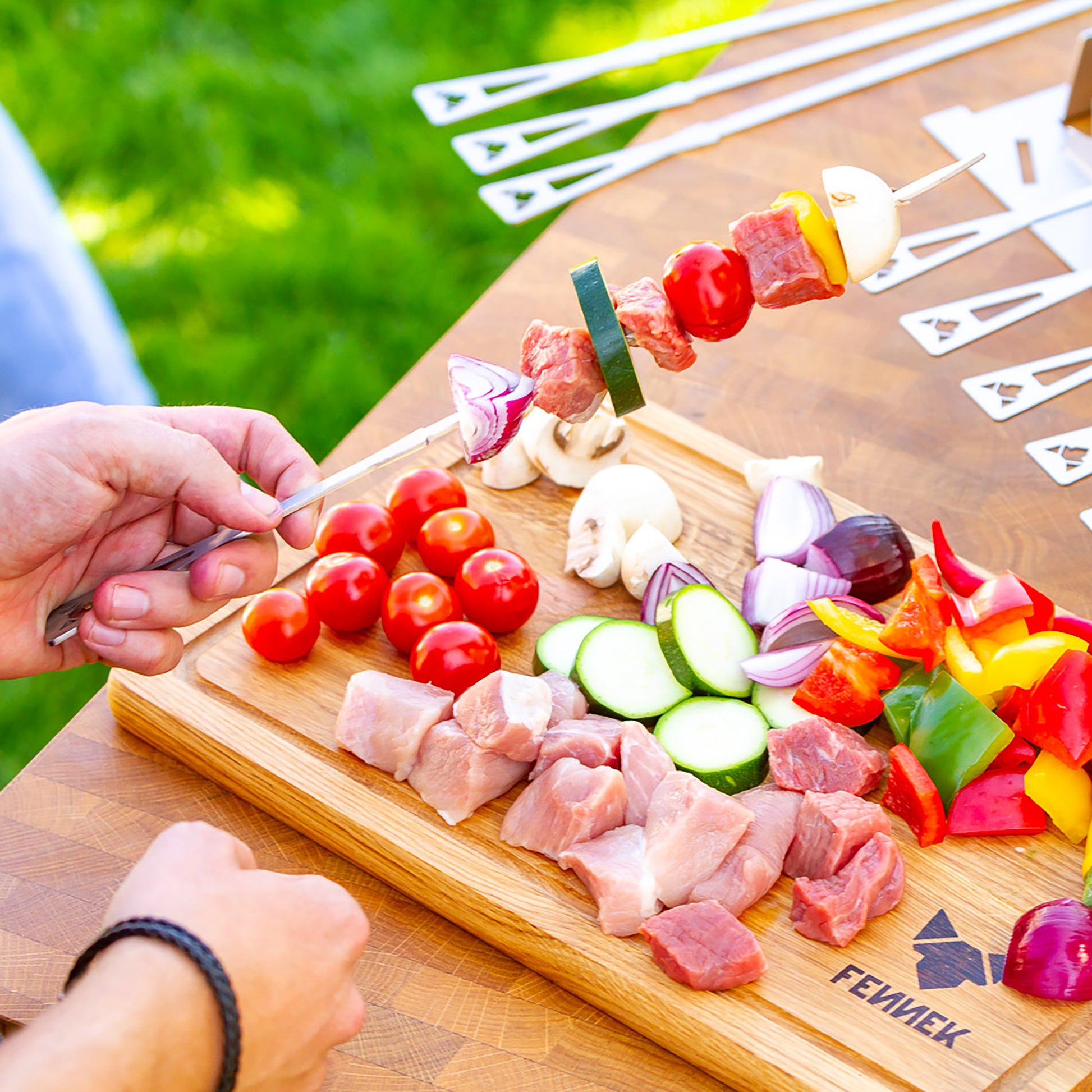 Auf einem Schneidebrett mit FENNEK Logo-Branding liegt kleingeschnittenes Fleisch und buntes Gemüse und wird auf Grillspieße aus Edelstahl gesteckt.