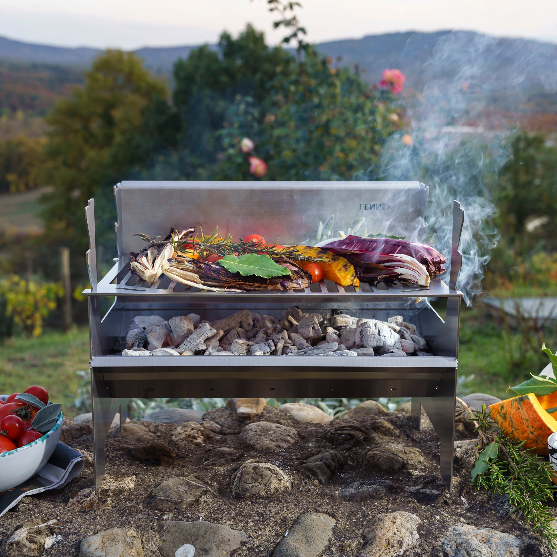 Fennek Holzkohlegrill 2.0 aus Edelstahl aufgebaut und mit brennender Kohle darin und Grillgut auf dem Rost steht im Grünen.