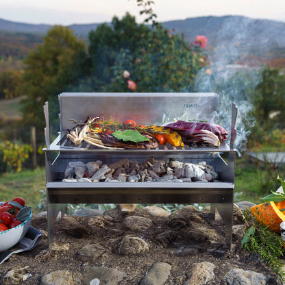 Fennek Holzkohlegrill 2.0 aus Edelstahl aufgebaut und mit brennender Kohle darin und Grillgut auf dem Rost steht im Grünen.