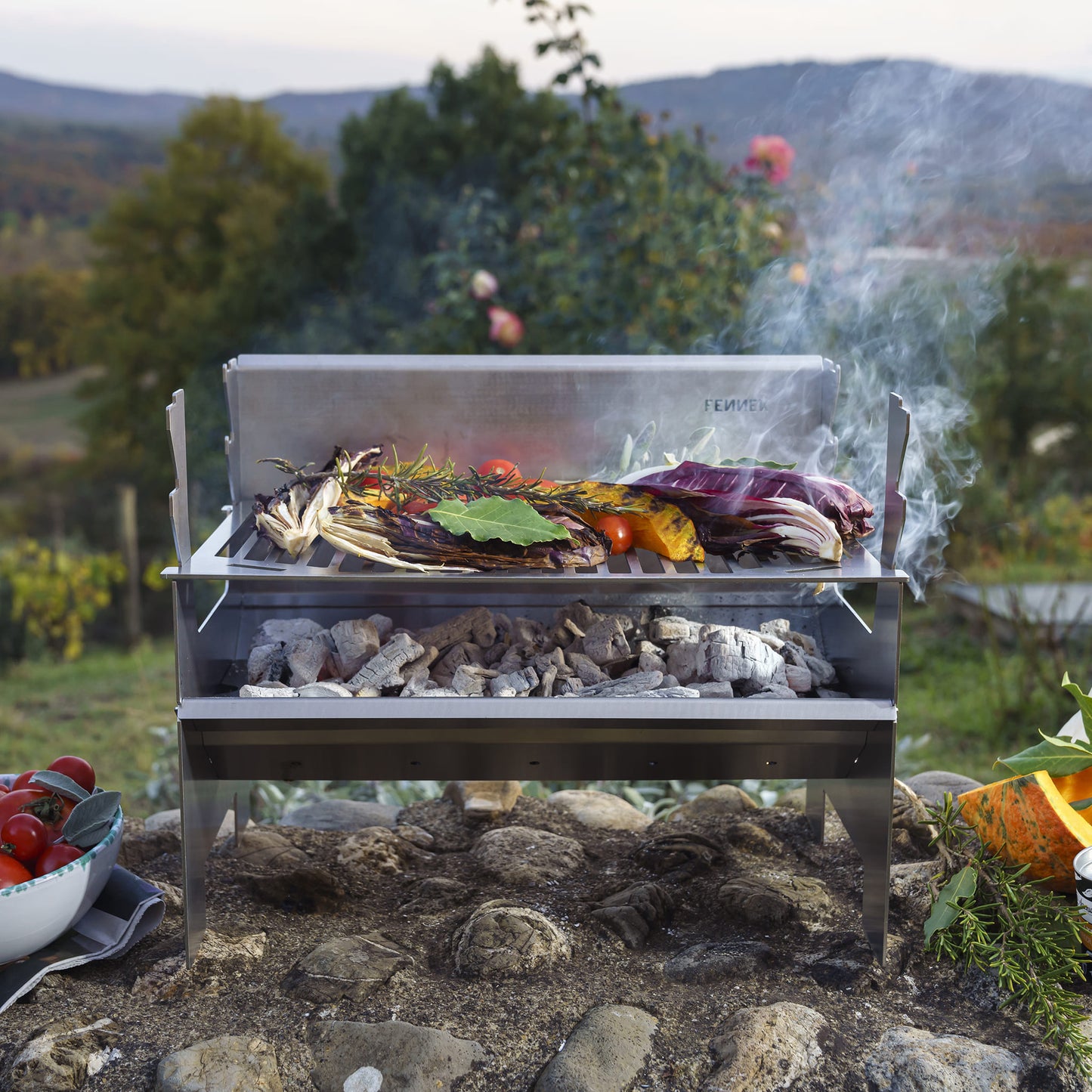 Fennek Holzkohlegrill 2.0 aus Edelstahl aufgebaut und mit brennender Kohle darin und Grillgut auf dem Rost steht im Grünen.