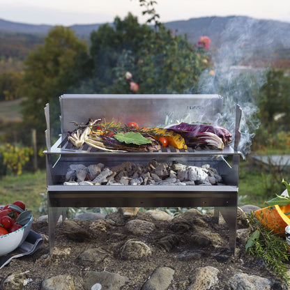 Fennek Holzkohlegrill 2.0 aus Edelstahl aufgebaut und mit brennender Kohle darin und Grillgut auf dem Rost steht im Grünen.