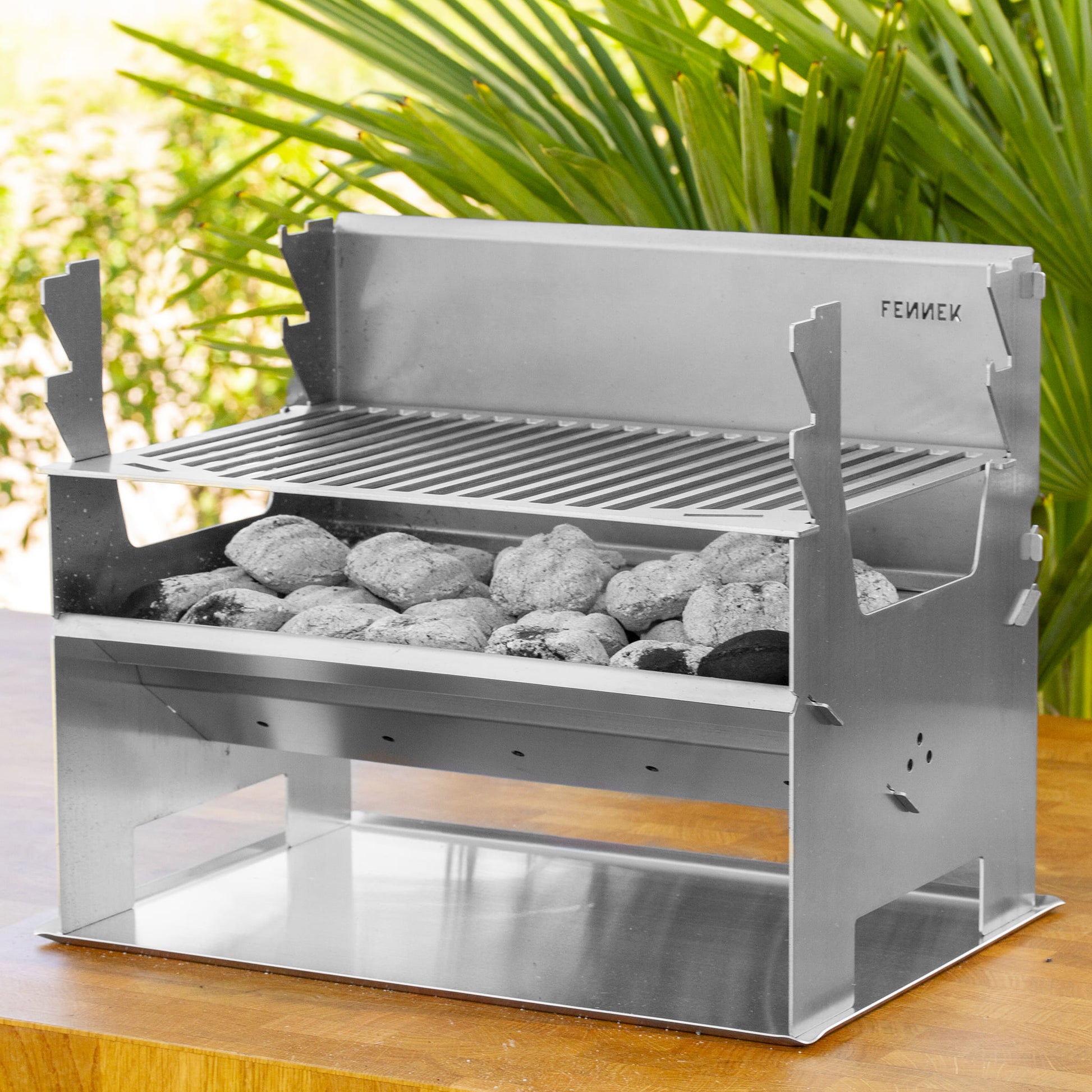 Fennek Holzkohlegrill 2.0 aus Edelstahl aufgebaut und mit brennender Kohle darin auf einem Bodenblech das auf einem Holztisch im Garten steht.