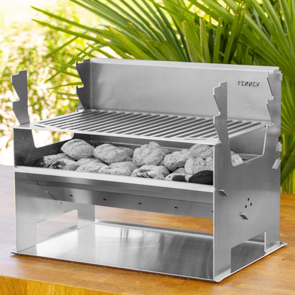 Fennek Holzkohlegrill 2.0 aus Edelstahl aufgebaut und mit brennender Kohle darin auf einem Bodenblech das auf einem Holztisch im Garten steht.