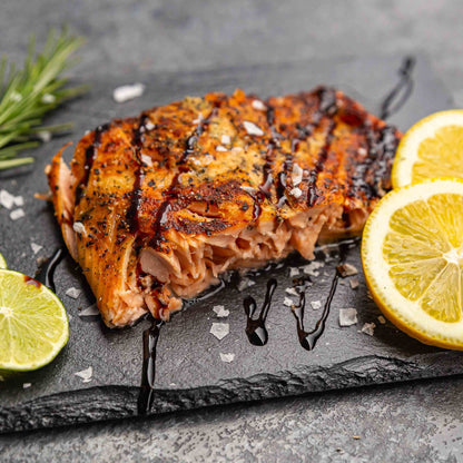 Zubereiteter Flammlachs lecker angerichtet auf einem schwarzen Schieferbrett mit Rosmarin, Limette und Zitrone dekoriert und Salzflocken und Balsamico gewürzt.