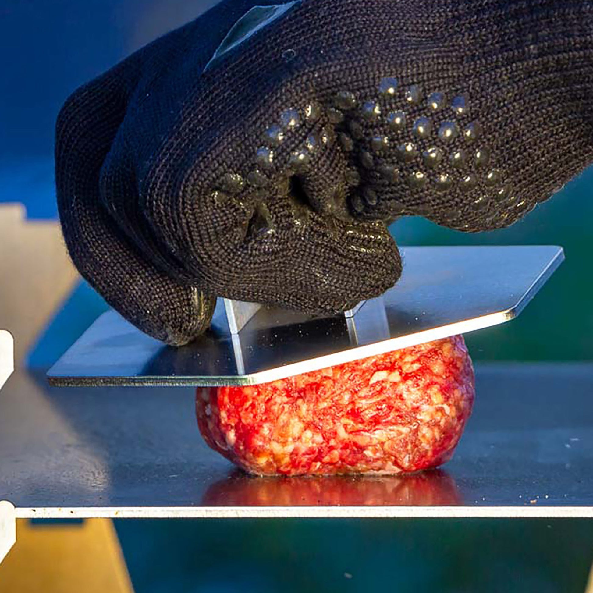 Schwarzer Grillhandschuh hält 6-eckige Hamburger Presse aus Edelstahl und presst einen Burger Patty auf der Plancha eines Holzkohlegrills.
