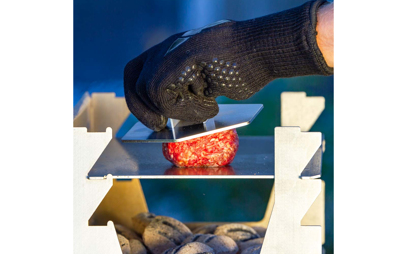 Schwarzer Grillhandschuh hält 6-eckige Hamburger Presse aus Edelstahl und presst einen Burger Patty auf der Plancha eines Holzkohlegrills.