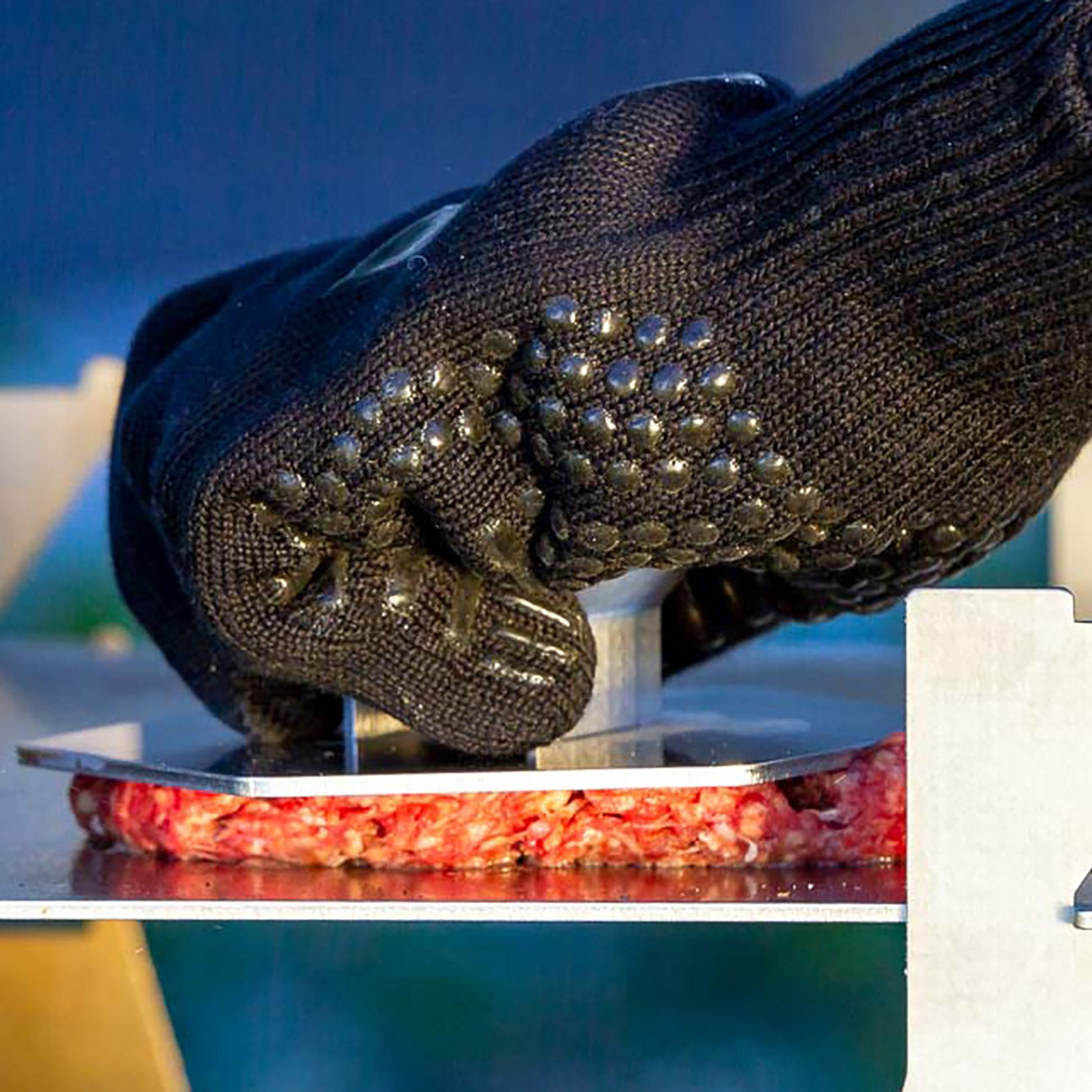 Schwarzer Grillhandschuh hält 6-eckige Hamburger Presse aus Edelstahl und presst einen Burger Patty auf der Plancha eines Holzkohlegrills.