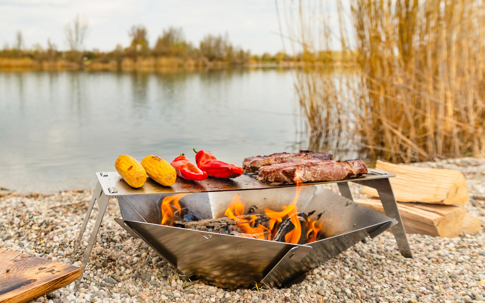 Die FENNEK Feuerschale Hexagon steht in einem Kiesbett am See. Darüber ist der FENNEK 4fire aufgestellt, auf welchem zwei Steaks und Gemüse gegrillt wird.