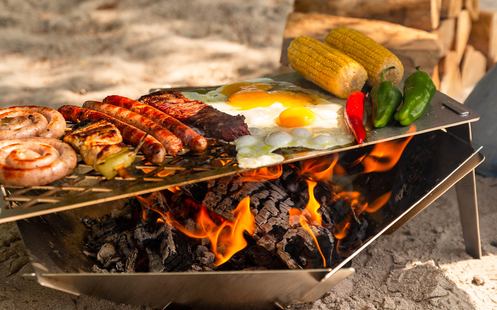 Fleisch und Gemüse wird auf einer zusammengesteckten Grillplatte über einer Feuerschale gegrillt die sich mit Erhöhung auf einem sandigen Untergrund