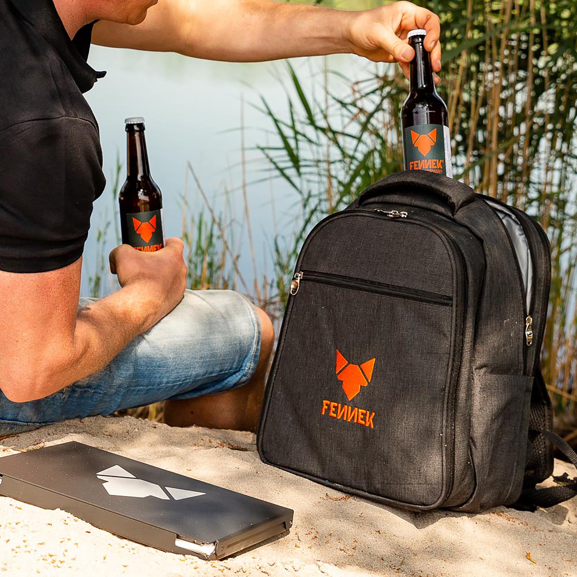 Rucksack mit FENNEK Logo-Applikation und FENNEK Grill in schwarzer Alubox liegen im Sand vor einem See und jemand holt 2 Flaschen aus dem Kühlfach.