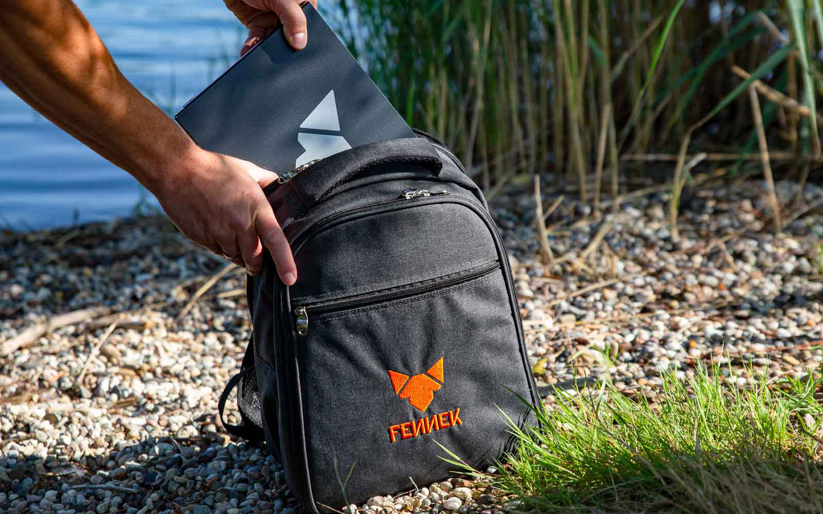Der Backpack steht auf einem Kiesbett. Im Hintergrund ist ein See zu sehen. Ein Mann packt den FENNEK Grill, welcher in seinem dazugehörigen Alukoffer eingepackt ist in den Backpack ein.