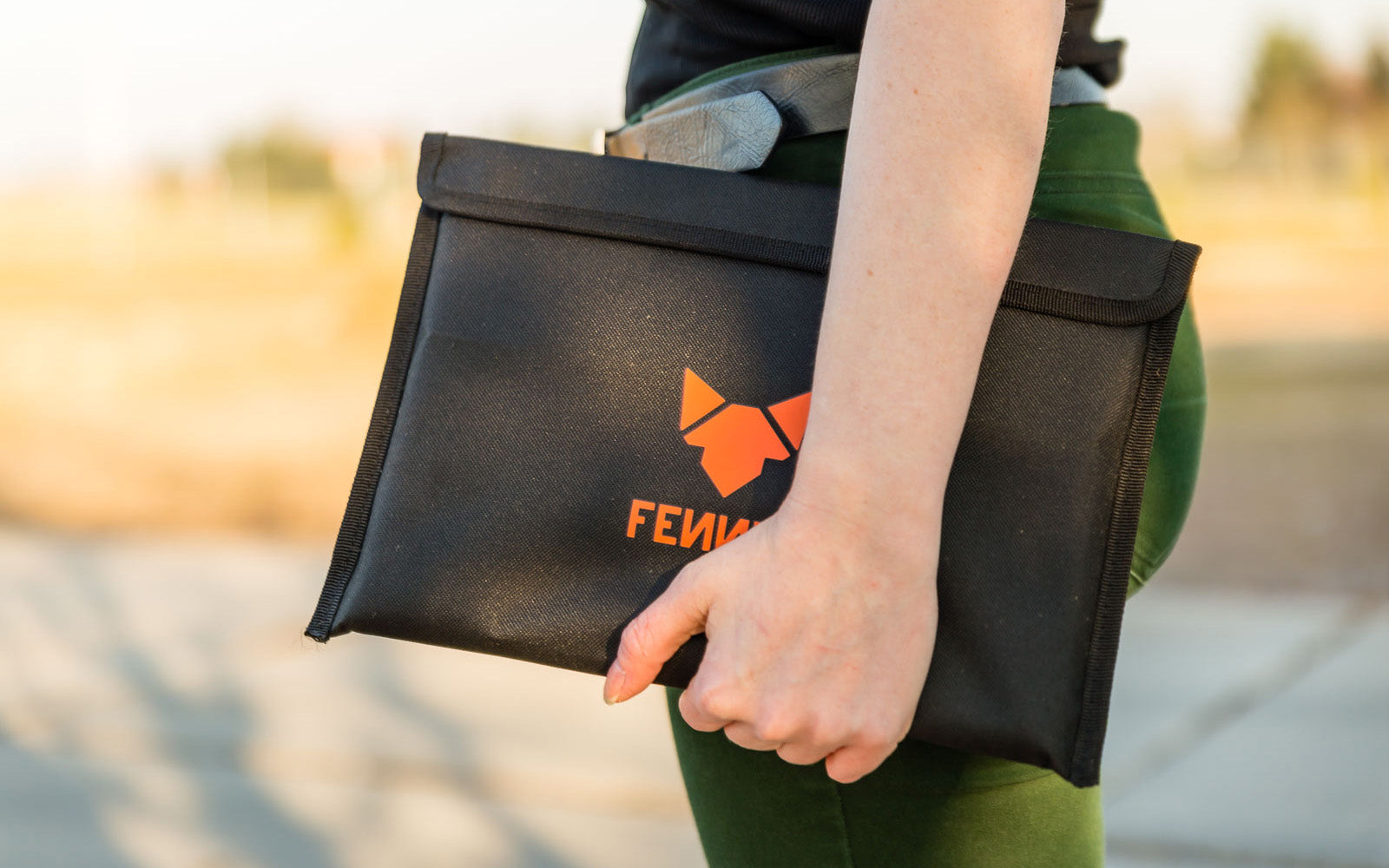 Eine Person trägt eine grüne Hose und hält auf Hüfthöhe eine schwarze Nylon-Tasche mit FENNEK Logo-Aufdruck