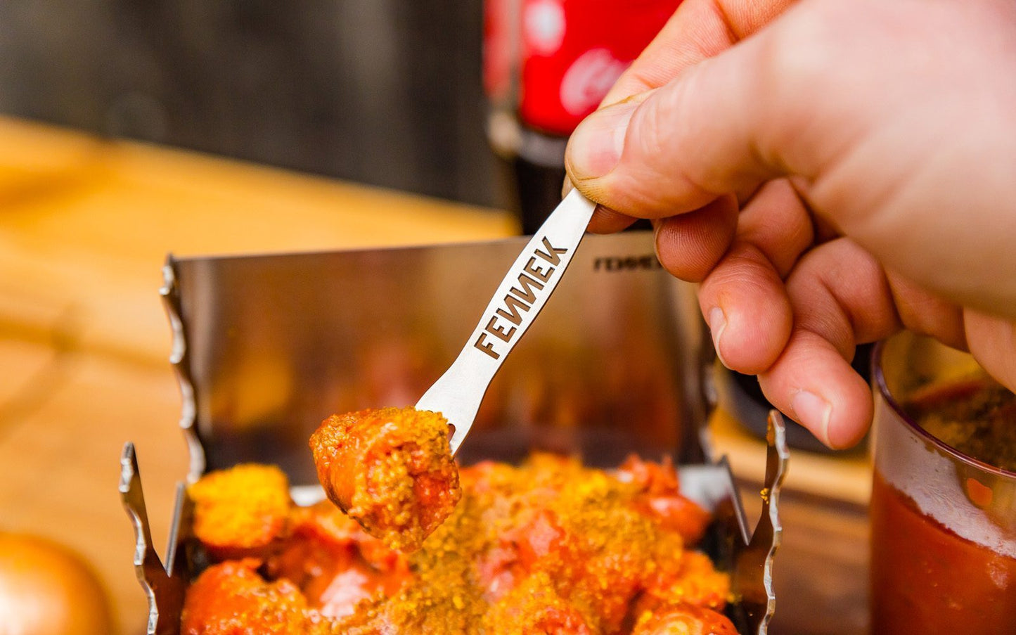Currywurst auf einer Edelstahl Pommesgabel mit herausgeschnittenem FENNEK Schriftzug in der Gabel