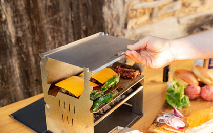 Eine Hand verstellt den Deckel eines Holzkohlegrills auf dem Cheeseburger zubereitet werden.