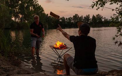 Ein Mann wirft in der Abenddämmerung einem anderen Brennholz zu. Brennendes Feuer im Feuernetz das nach Sonnenuntergang im See steht.