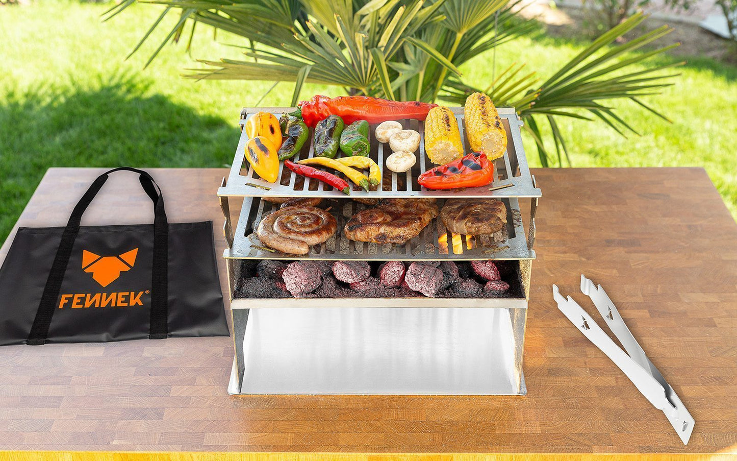 FENNEK 2.0 Holzkohlegrill aus Edelstahl mit 2. Grillrost und viel buntem Gemüse und Grillfleische auf einem Holztisch mit Tasche und Grillzange daneben im Grünen Garten.