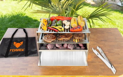 FENNEK 2.0 Holzkohlegrill aus Edelstahl mit 2. Grillrost und viel buntem Gemüse und Grillfleische auf einem Holztisch mit Tasche und Grillzange daneben im Grünen Garten.