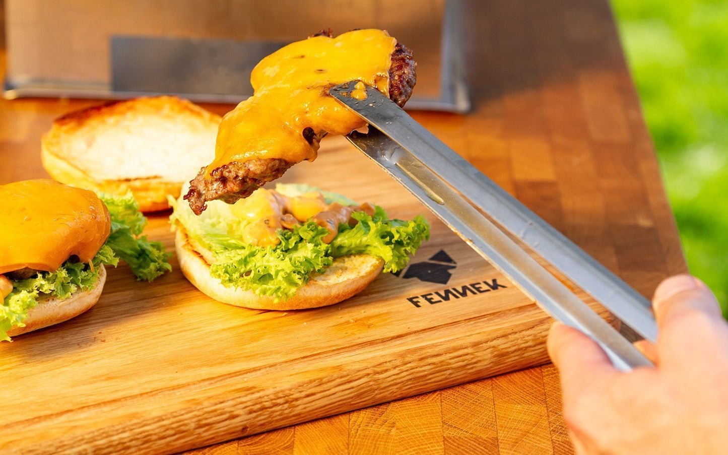 Mit Käse überbackenes Hamburger Patty wird mit Grillzange aus Edelstahl auf ein Brötchen gelegt, darunter ein Holzbrett mit Fennek Logo-Branding.