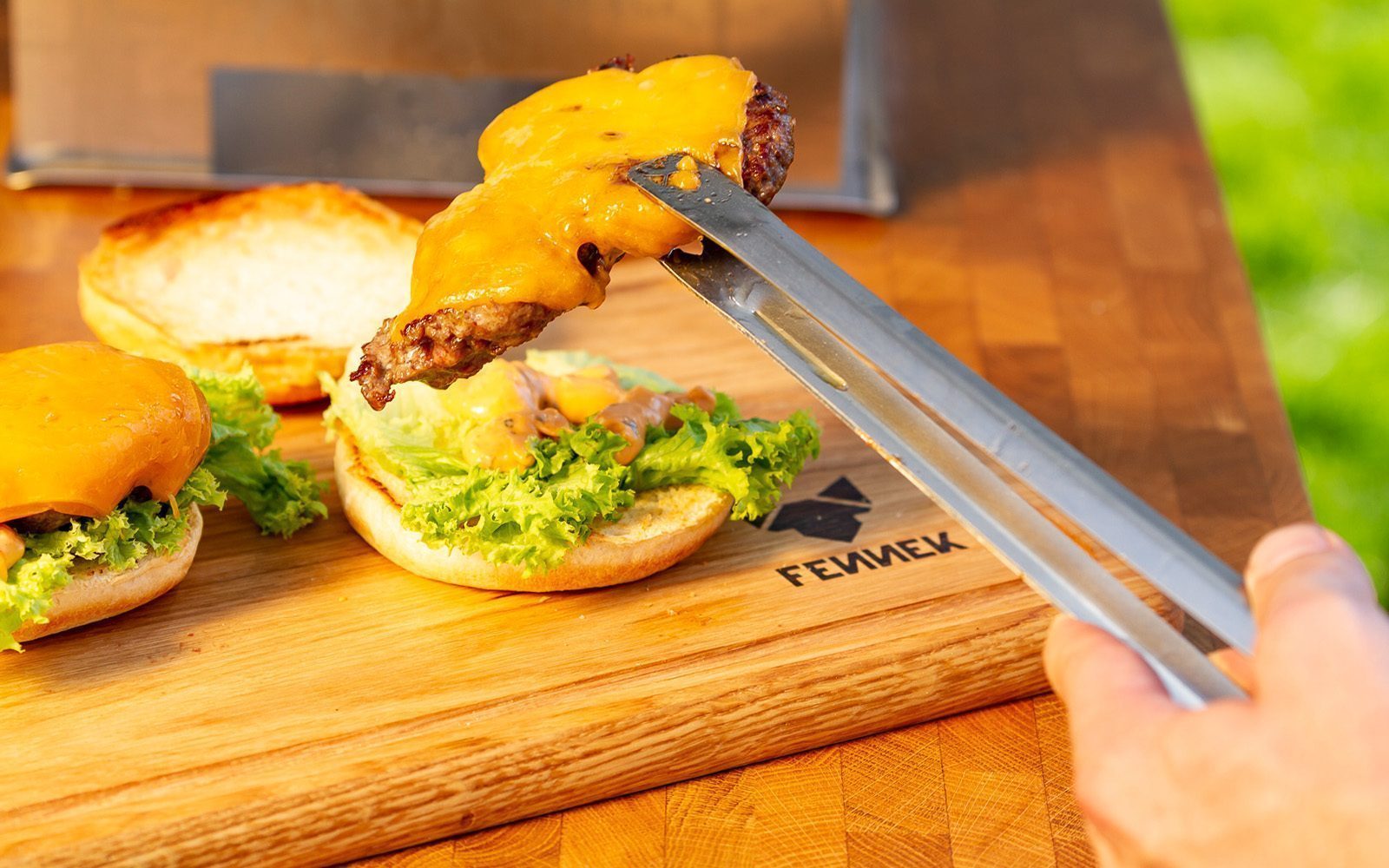 Mit Käse überbackenes Hamburger Patty wird mit Grillzange aus Edelstahl auf ein Brötchen gelegt, darunter ein Holzbrett mit Fennek Logo-Branding.